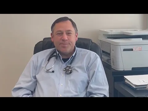 Dr. Steve Johnson, in a light blue shirt and a stethoscope, sits at his desk beside a printer.