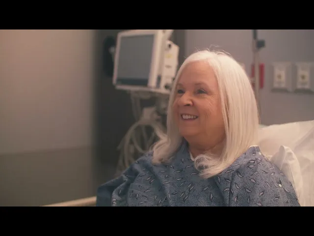 El video aún está en pausa y muestra a una mujer sonriente con una bata de hospital azul en una cama de hospital.